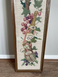 Broderie néerlandaise encadrée représentant une vigne avec des raisins, des bouteilles de vin et des paniers.