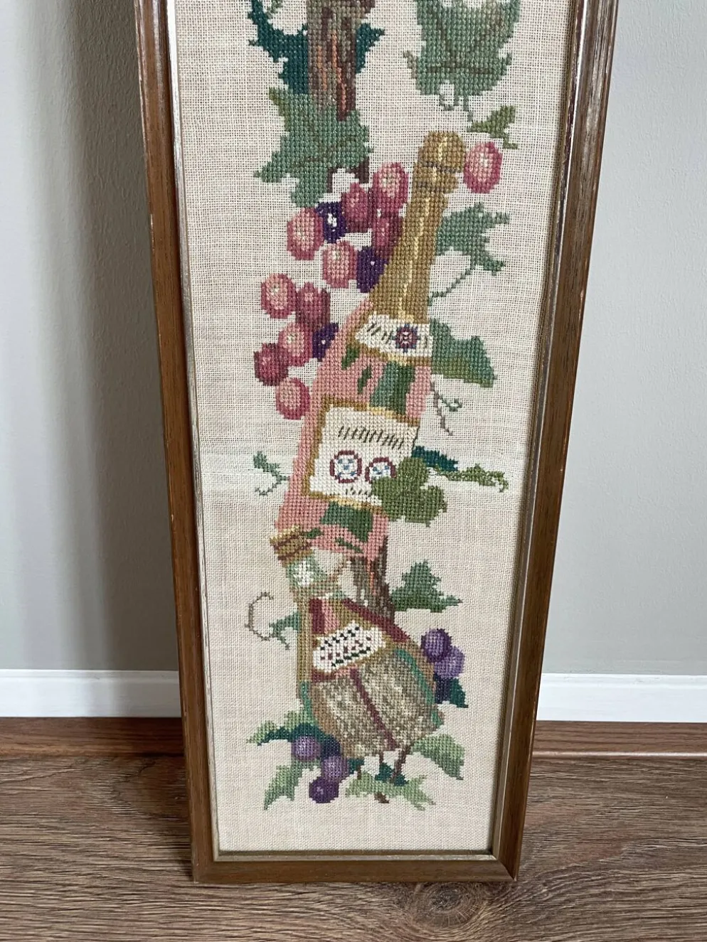 Broderie néerlandaise encadrée représentant une vigne avec des raisins, des bouteilles de vin et des paniers.
