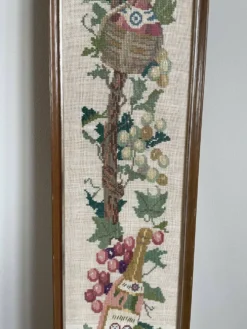 Broderie néerlandaise encadrée représentant une vigne avec des raisins, des bouteilles de vin et des paniers.