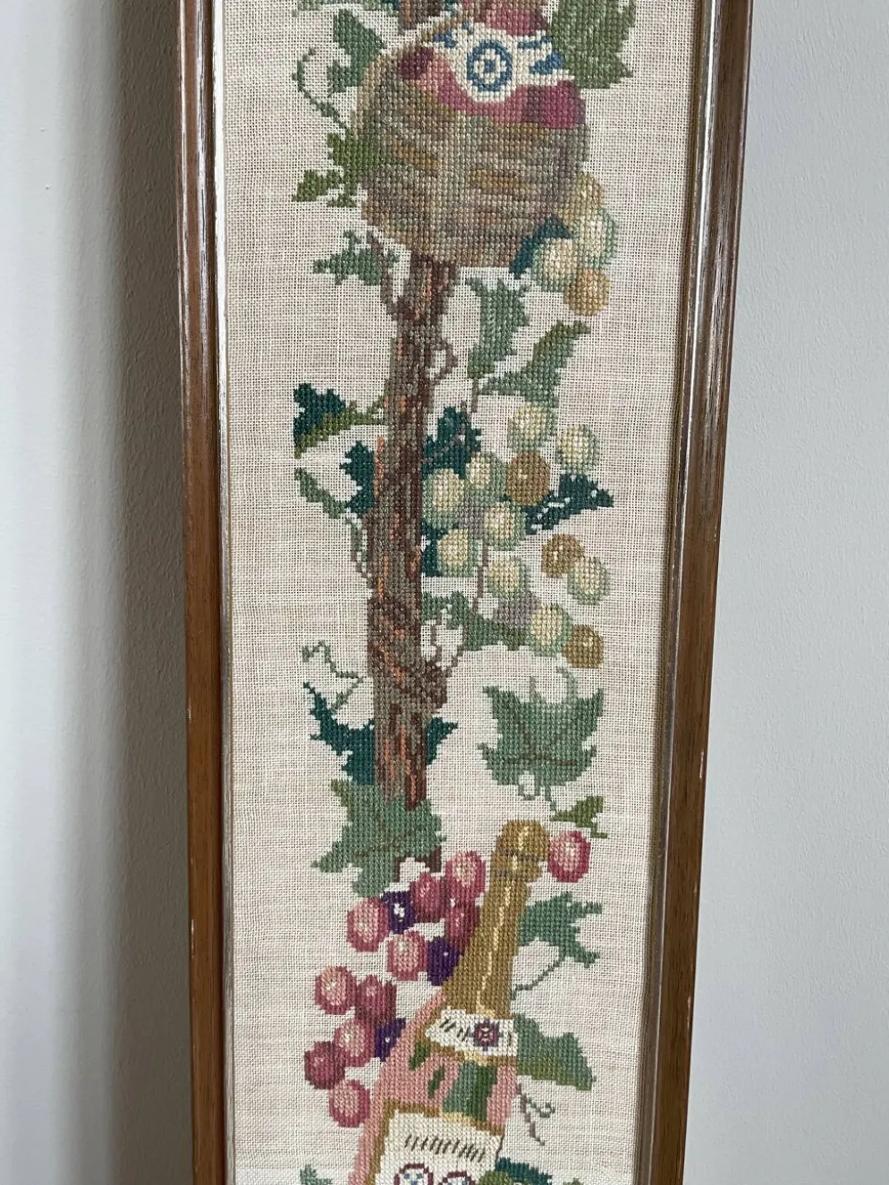Broderie néerlandaise encadrée représentant une vigne avec des raisins, des bouteilles de vin et des paniers.