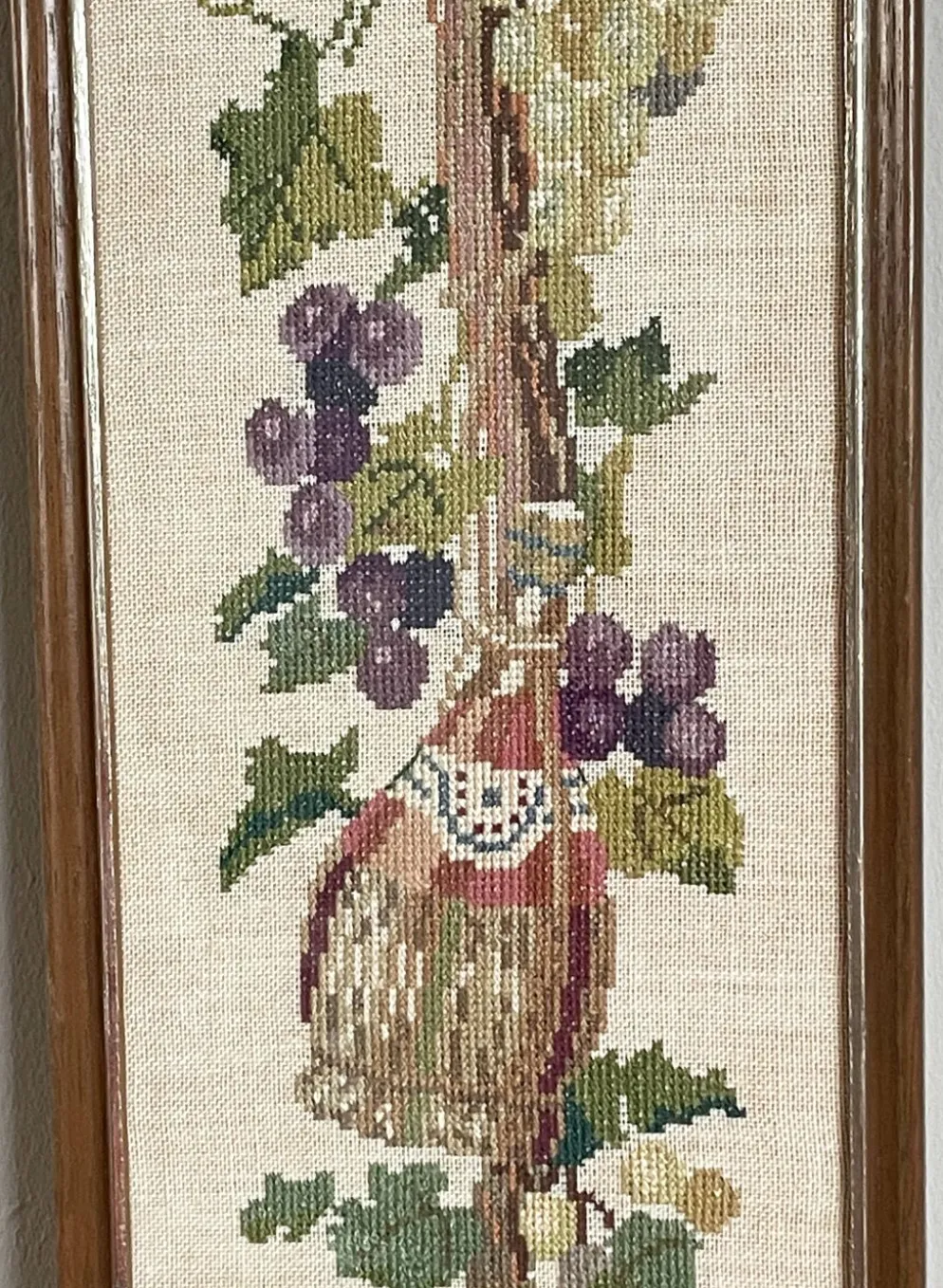 Broderie néerlandaise encadrée représentant une vigne avec des raisins, des bouteilles de vin et des paniers.
