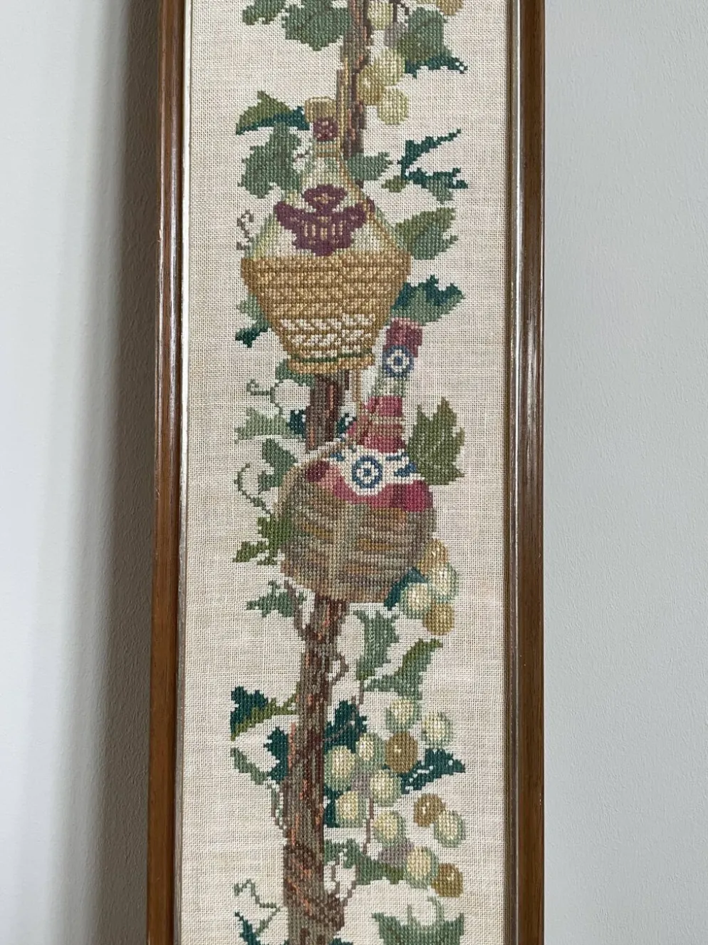 Broderie néerlandaise encadrée représentant une vigne avec des raisins, des bouteilles de vin et des paniers.