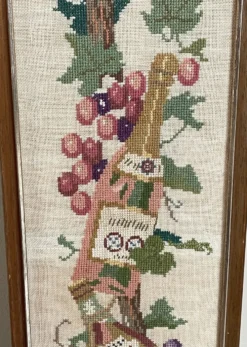 Broderie néerlandaise encadrée représentant une vigne avec des raisins, des bouteilles de vin et des paniers.