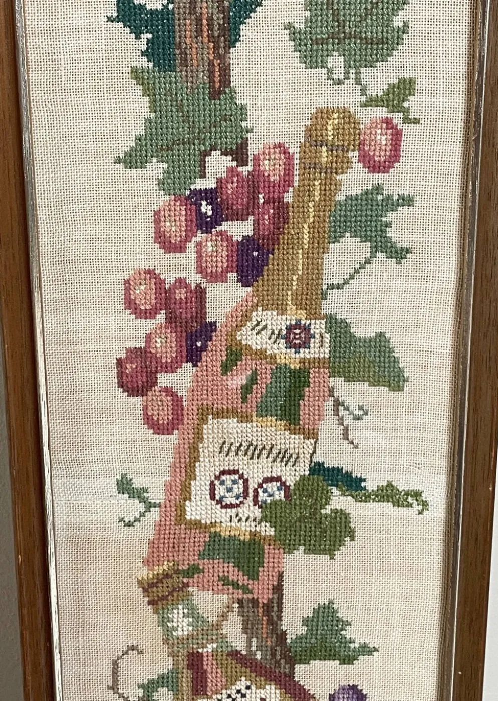 Broderie néerlandaise encadrée représentant une vigne avec des raisins, des bouteilles de vin et des paniers.