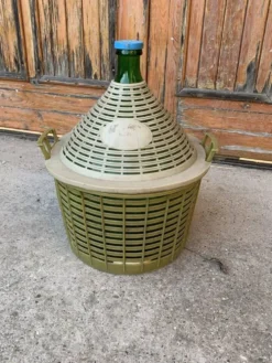 Grande dame‑jeanne en verre vert dans panier plastique kaki style mid‑century